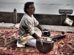 A picture of a man sitting with his legs crossed on a rug. He has a sitar in front of him.