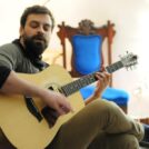 Jordan Zalis, a man playing an acoustic guitar. Behind him is a blue chair.