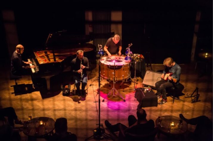 A group of four musicians playing on a stage. The instruments they are using are a piano, a clarinet, a large drum, and an electric guitar.