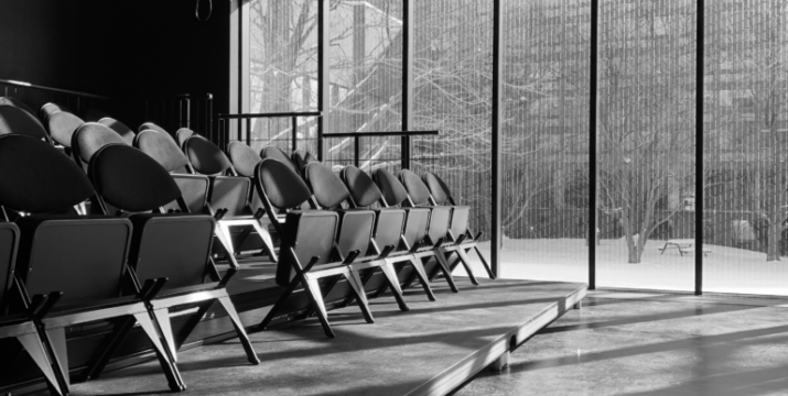A black and white of the interior of ImprovLab. Raised theatre seating sits in front of large vertical glass windows.