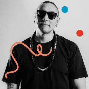 A portrait of Classic Roots wearing a black shirt, sunglasses and a necklace, standing against an off-white background. Image has been filtered in greyscale with colourful dots and a squiggly line added.