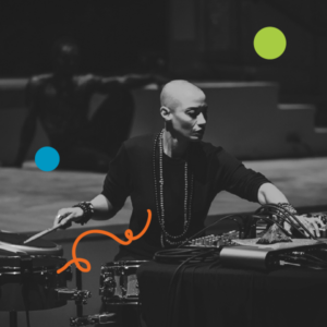 A photo of Val Jeanty performing on stage with acoustic and electric drums, as well as laptops. Behind her is a seated dancer. Image is filtered in greyscale with colourful dots and a squiggly line added.