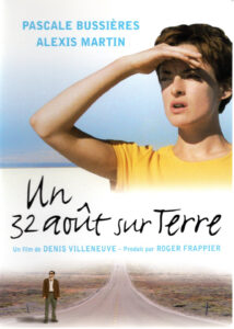 Poster for Un 32 août sur terre. The poster is divided in half by a white background with the stylized title text, director credit, and producer credit. The top half of the poster features a person with short hair in a yellow t-shirt shielding their eyes against the sun, with blue sky in the background. The bottom portion of the image shows another person in a suit standing at the side of a long desert road stretching off towards a vanishing point in the centre of the poster.