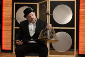 A photo of Jesse Stewart in a suit and bowler hat, seated, and playing a waterphone. The background features various large metallic discs arranged in frames.
