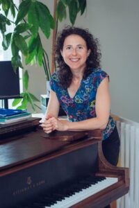 A photo of Marianne Trudel smiling and leaning forwards with her forearms resting on a piano.