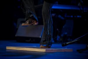 A photo of Heather Cornell's legs in dark pants with one leg raised as she dances against a black stage background with overhead blue lighting. Photo by William Frederking.